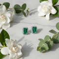 Emerald green CZ emerald-cut rectangle halo stud earrings on white marble with eucalyptus and gardenia - lifestyle style