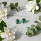 Emerald green CZ emerald-cut rectangle halo stud earrings on white marble with eucalyptus and gardenia - lifestyle style