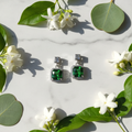 Emerald green CZ cushion halo drop earrings on white marble with eucalyptus and jasmine - lifestyle style
