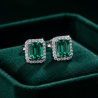 Emerald green CZ emerald-cut rectangle halo stud earrings on forest green velvet - dramatic editorial style