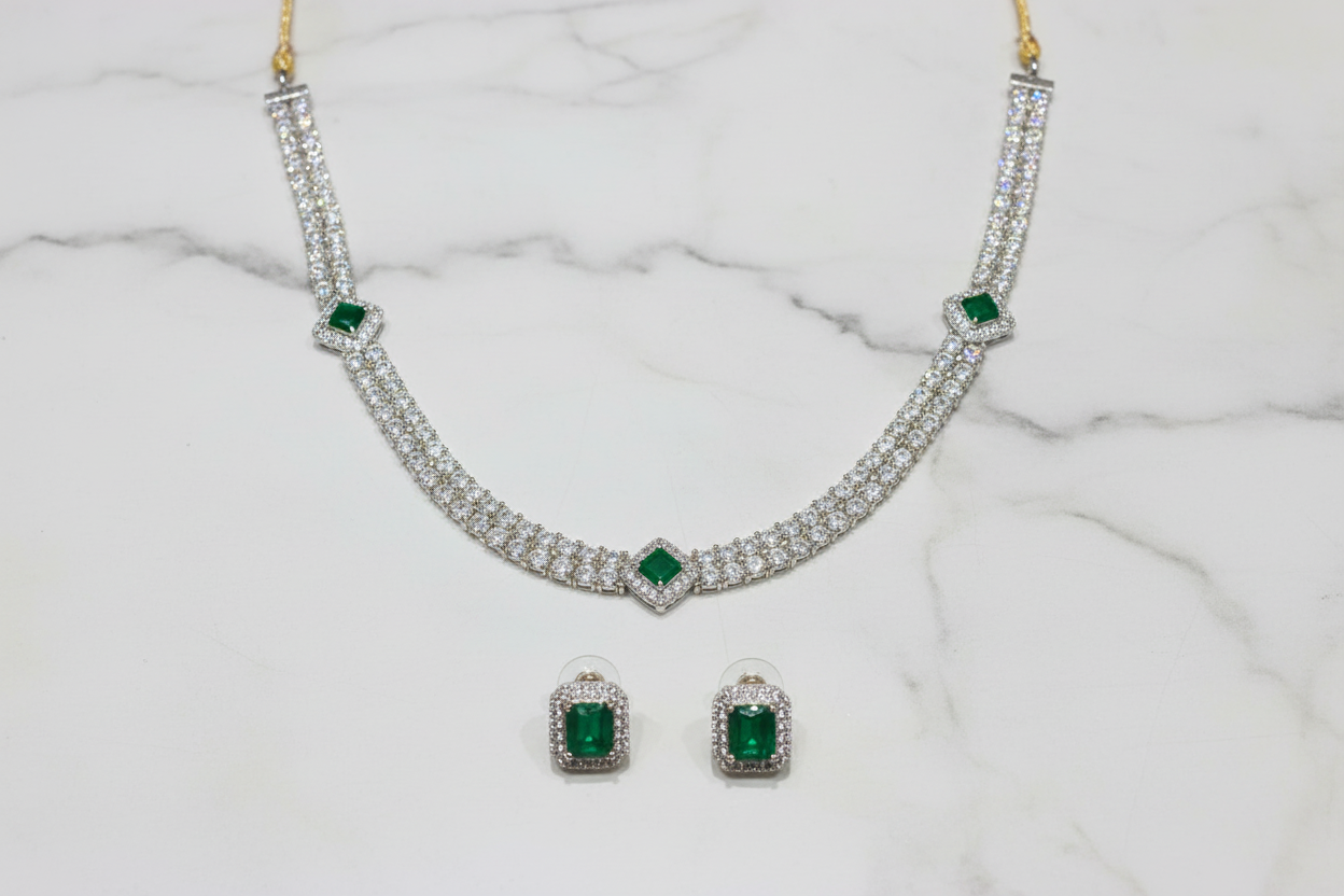 Flat lay of white AD tennis necklace with emerald stations and earrings on white marble