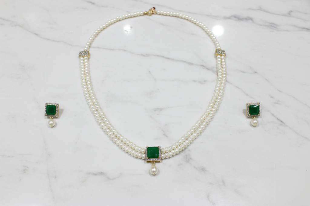 Flat lay of 2-strand seed pearl choker with emerald square AD pendant and earrings on white marble