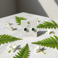 Emerald green CZ cushion square solitaire stud earrings on white marble with fern and jasmine - lifestyle style