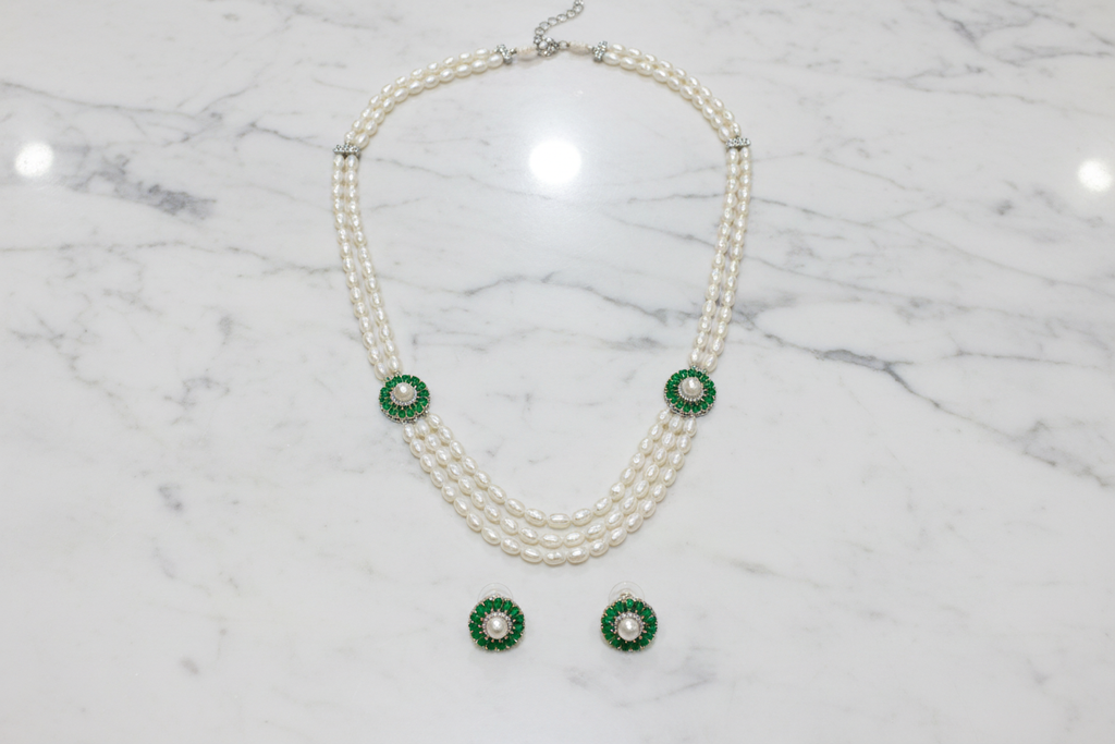 Flat lay of 3-strand pearl long necklace with emerald floral AD stations and earrings on white marble