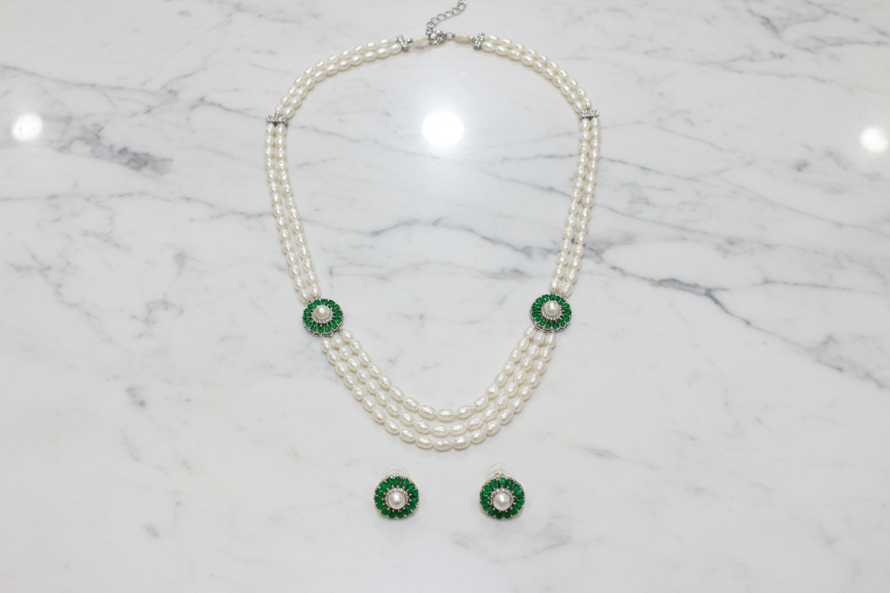 Flat lay of 3-strand pearl long necklace with emerald floral AD stations and earrings on white marble