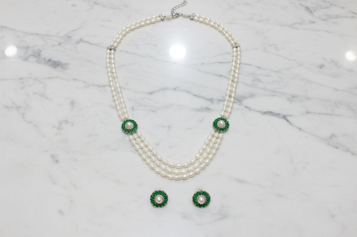 Flat lay of 3-strand pearl long necklace with emerald floral AD stations and earrings on white marble